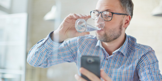 Hombre toma un vaso de agua mientras mira su celular